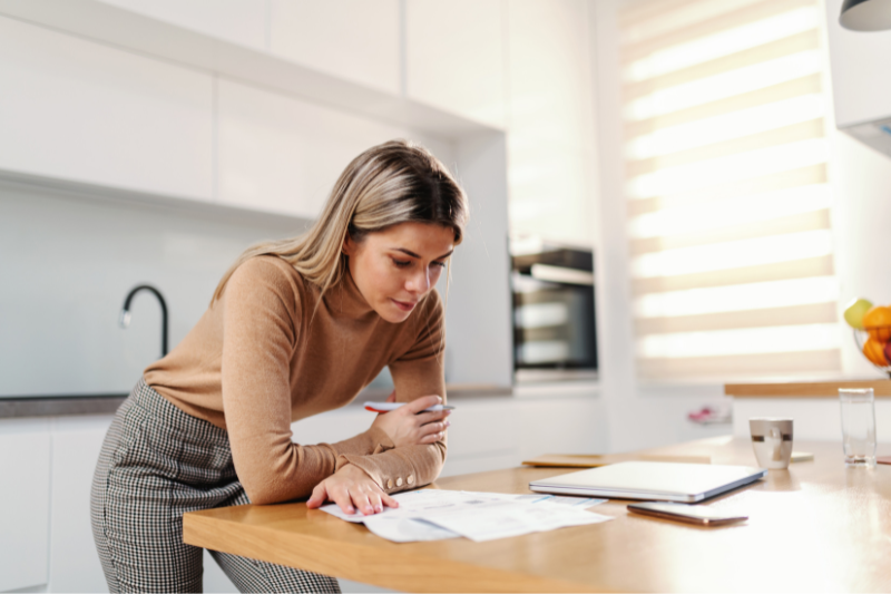A woman stands in a bright kitchen, leaning over a table while reviewing papers. A laptop, notebook, and pen are on the table, and she appears focused and thoughtful as she reads.